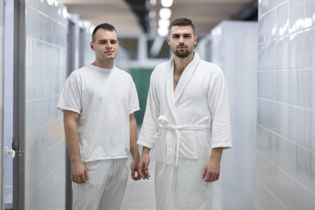 Medical Consultation in Spa Corridor.Two men discussing treatment options while walking through a clinical spa corridor, wearing white uniforms and bathrobe.の写真素材