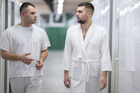Medical Consultation in Spa Corridor.Two men discussing treatment options while walking through a clinical spa corridor, wearing white uniforms and bathrobe.の写真素材