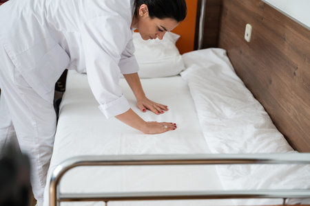 Two nurses in uniforms cleaning and organizing a hospital room by changing the bed linens and...