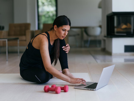 A young woman in sportswear is sitting in the living room and preparing for online training while using a laptop.の写真素材