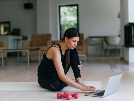 A young woman in sportswear is sitting in the living room and preparing for online training while using a laptop.の写真素材