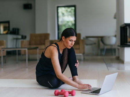 A young woman in sportswear is sitting in the living room and preparing for online training while using a laptop.の写真素材