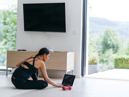 A young woman in sportswear is sitting in the living room and preparing for online training while using a laptop.の写真素材