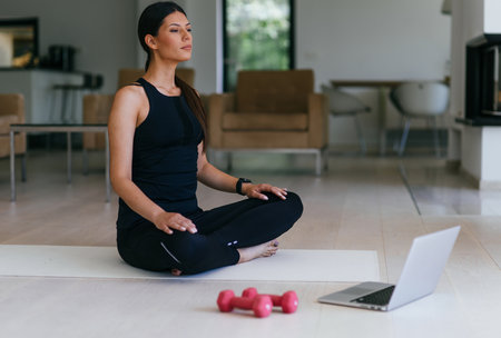 A young woman in sportswear is sitting in the living room and preparing for online training while using a laptop.の写真素材