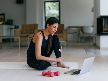 A young woman in sportswear is sitting in the living room and preparing for online training while using a laptop.の写真素材