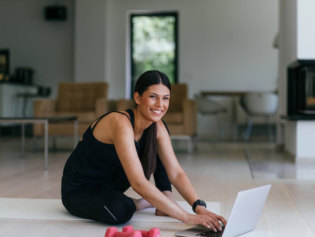 A young woman in sportswear is sitting in the living room and preparing for online training while using a laptop.の写真素材