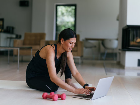 A young woman in sportswear is sitting in the living room and preparing for online training while using a laptop.の写真素材