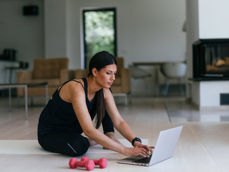 A young woman in sportswear is sitting in the living room and preparing for online training while using a laptop.の写真素材