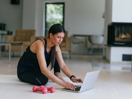 A young woman in sportswear is sitting in the living room and preparing for online training while using a laptop.の写真素材
