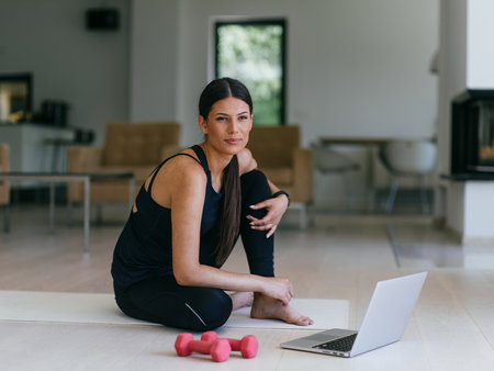 A young woman in sportswear is sitting in the living room and preparing for online training while using a laptop.の写真素材