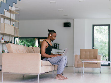 African American man in glasses sitting at a table in a modern living room, using a laptop for business video chat, conversation with friends and entertainment.の写真素材