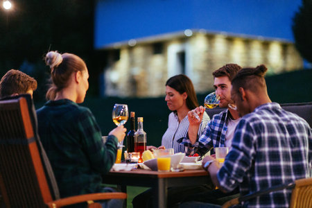A group of young diverse people having dinner on the terrace of a modern house in the evening. Fun for friends and family. Celebration of holidays, weddings with barbecueの写真素材