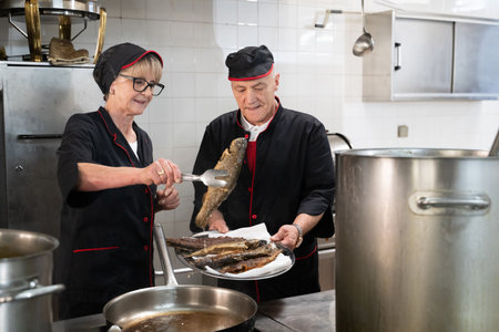 Two chefs in black uniforms working together while serving fish in a commercial kitchen. Concept of seafood preparation, teamwork, culinary skills, and hospitality industry.の写真素材