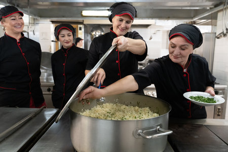Group of chefs in black uniforms preparing rice in a large pot in a commercial kitchen. Female cooks stirring and adding herbs while working as a team. Concept of teamwork, gastronomy, and hospitality industry.の写真素材