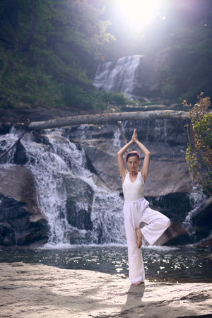 Woman doing yoga tree pose in front of waterfall at sunrise peaceful meditation in natureの写真素材