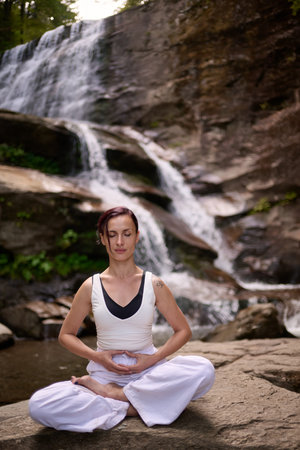 Young woman sitting in lotus pose under tropical waterfall meditating with calm strength and mindfulness fully connected to nature energy tranquility and inner balanceの写真素材