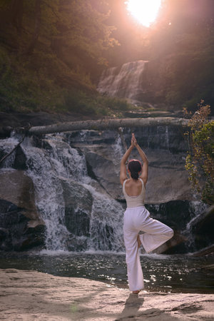 Woman doing yoga tree pose in front of waterfall at sunrise peaceful meditation in natureの写真素材