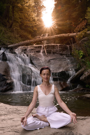 Young woman sitting in lotus pose under tropical waterfall meditating with calm strength and mindfulness fully connected to nature energy tranquility and inner balanceの写真素材