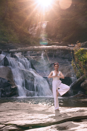 Woman doing yoga tree pose in front of waterfall at sunrise peaceful meditation in natureの写真素材