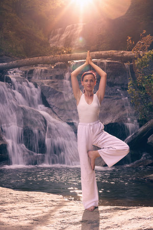 Woman doing yoga tree pose in front of waterfall at sunrise peaceful meditation in natureの写真素材