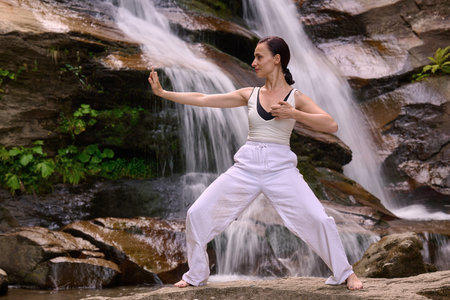 woman performing slow tai chi movements near a waterfall and river at sunset capturing harmony balance and fitness in natureの写真素材
