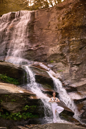 Woman doing yoga tree pose in front of waterfall at sunrise peaceful meditation in natureの写真素材