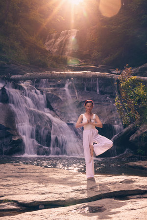 Woman doing yoga tree pose in front of waterfall at sunrise peaceful meditation in natureの写真素材