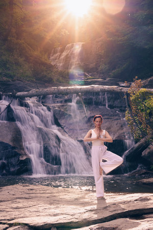Woman doing yoga tree pose in front of waterfall at sunrise peaceful meditation in natureの写真素材