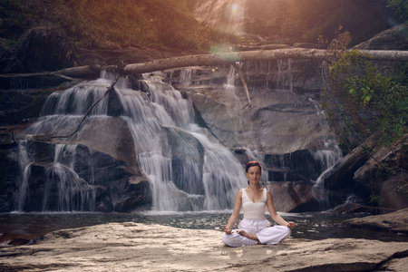 Young woman sitting in lotus pose under tropical waterfall meditating with calm strength and mindfulness fully connected to nature energy tranquility and inner balanceの写真素材