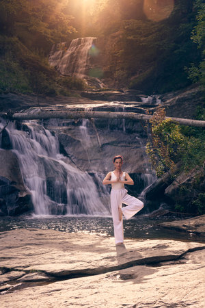 Woman doing yoga tree pose in front of waterfall at sunrise peaceful meditation in natureの写真素材