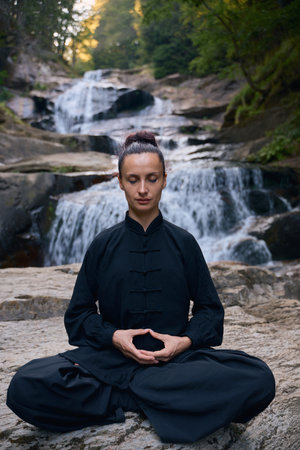 A woman sits in lotus pose, practicing yoga and tai chi under a tropical waterfall, meditating with calm strength and mindfulness, fully connected to the energy of nature, tranquility and inner balance.の写真素材