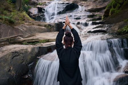 Woman practicing Tai Chi in nature, wearing a traditional black martial arts uniform, standing gracefully near a waterfall, focused on meditation and balance. Healthy lifestyle in asian cultureの写真素材