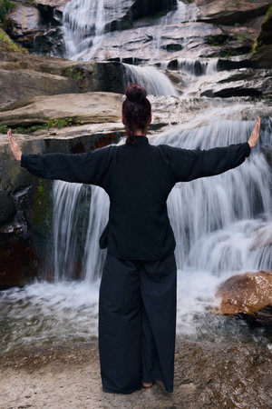 Woman practicing Tai Chi in nature, wearing a traditional black martial arts uniform, standing gracefully near a waterfall, focused on meditation and balance. Healthy lifestyle in asian cultureの写真素材