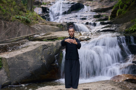 Woman practicing Tai Chi in nature, wearing a traditional black martial arts uniform, standing gracefully near a waterfall, focused on meditation and balance. Healthy lifestyle in asian cultureの写真素材
