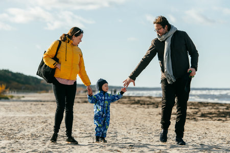 A happy family strolls along a chilly beach, with their young son joyfully running ahead, creating...