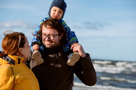 A happy family strolls along a chilly autumn beach, with their young son joyfully running ahead,...