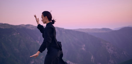 Woman practicing Tai Chi in traditional black outfit on mountain at sunrise, arms outstretched in...