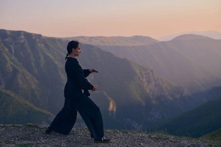 Woman practicing Tai Chi in traditional black outfit on mountain at sunrise, arms outstretched in...