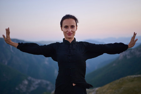 Woman practicing Tai Chi in traditional black outfit on mountain at sunrise, arms outstretched in...