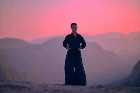 Woman practicing Tai Chi in traditional black outfit on mountain at sunrise, arms outstretched in...