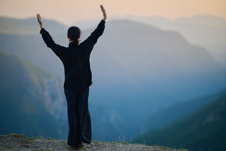 Woman practicing Tai Chi in traditional black outfit on mountain at sunrise, arms outstretched in...