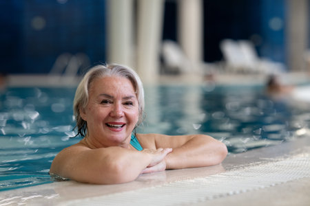 Smiling senior woman enjoying relaxation in an indoor swimming pool, leaning on the poolside. Concept of active lifestyle, wellness, health, hydrotherapy, and leisure for older adults.の写真素材