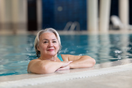 Smiling senior woman enjoying relaxation in an indoor swimming pool, leaning on the poolside....