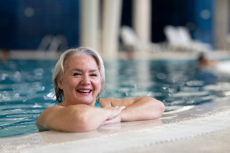 Smiling senior woman enjoying relaxation in an indoor swimming pool, leaning on the poolside. Concept of active lifestyle, wellness, health, hydrotherapy, and leisure for older adults.の写真素材