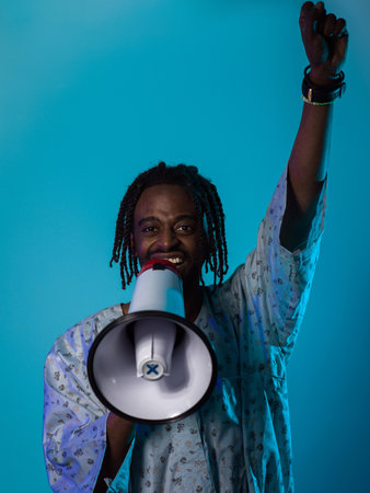 In a powerful and symbolic image, an African American man wears traditional clothing, passionately wields a megaphone against a striking blue background, with his hand raised in the air symbolizing his vocal and cultural empowerment in the pursuit of social justice and equalityの写真素材