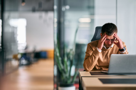 Concentrated businessman sitting at desk using laptop in modern office interior, thinking and analyzing data or business strategy, symbolizing productivity, focus, and professional work.の写真素材
