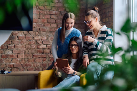 Three cheerful young women sitting in a cozy modern interior, smiling and looking at a tablet screen together, representing friendship, technology, and digital lifestyle.の写真素材
