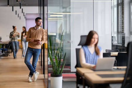 Businessman standing by glass wall in a modern office corridor, holding a digital tablet and smiling with reflection visible on the glass, symbolizing success, optimism, and modern business lifestyle.の写真素材