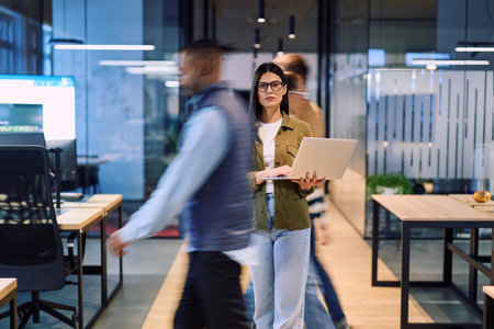 Business woman holding laptop in the middle of a dynamic modern office while colleagues move around, symbolizing focus, leadership, and productivity amid a fast-paced work environment.の写真素材