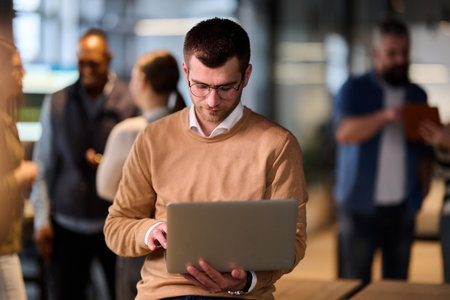Concentrated male professional using a laptop while standing in a modern open office space, with coworkers collaborating in the background, symbolizing productivity, focus, and teamwork.の写真素材